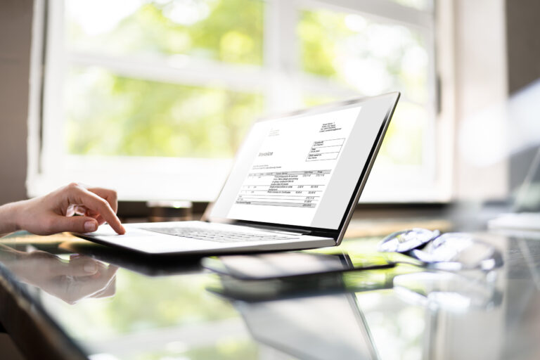 Person typing on a laptop displaying an invoice on the screen, with a bright window in the background.