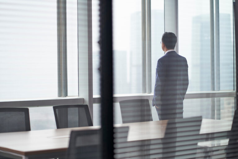rear side view of an asian business man standing in front of office window looking out and thinking hands in pocket